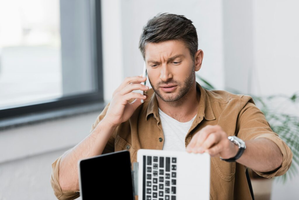 Skeptical businessman looking at broken laptop while talking on mobile phone on blurred background